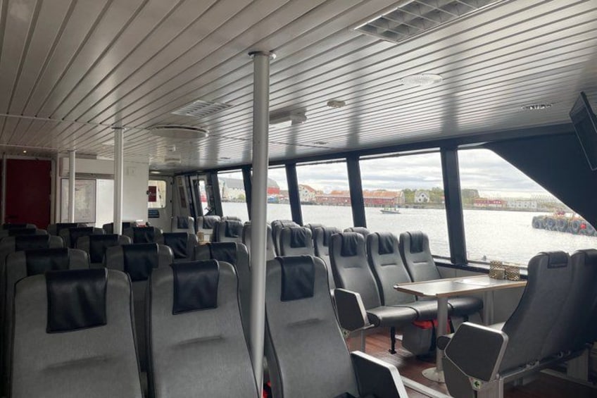 A ferry interior with rows of seats and tables, large windows, and a view of the water and cityscape. 3
