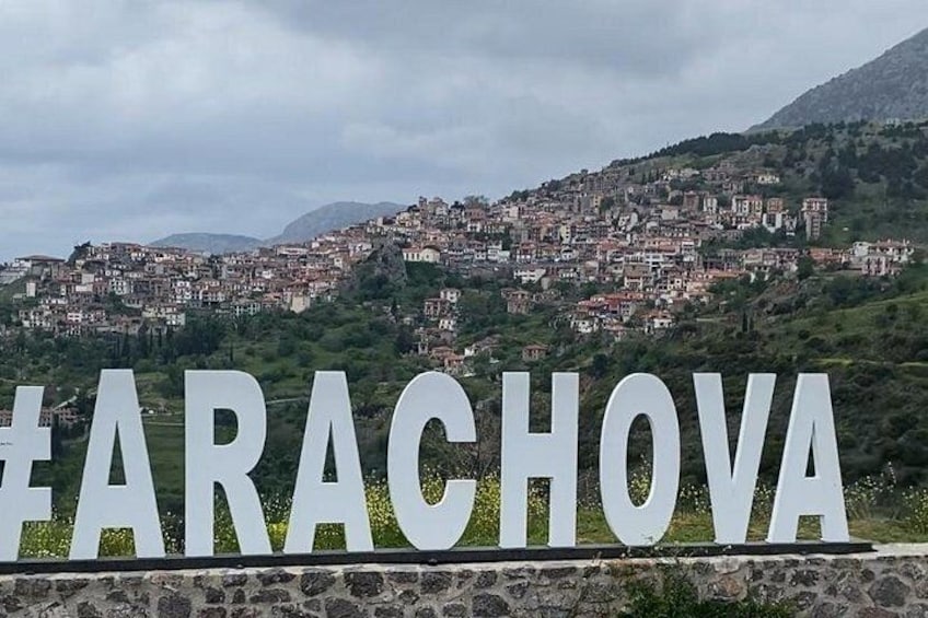 Ein Schild mit dem Wort 'ARACHOVA' in großen Buchstaben vor dem Hintergrund einer hügeligen Stadt mit Gebäuden. 4