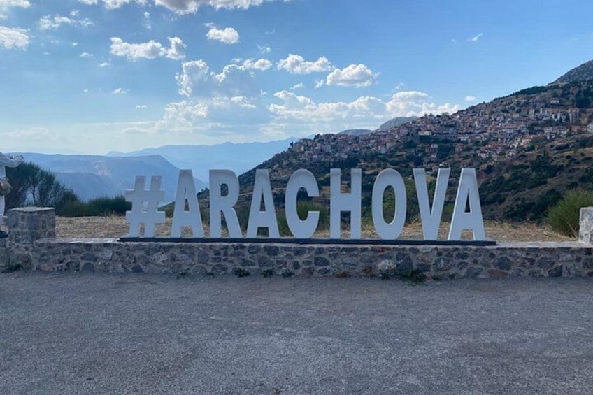 Ein Schild mit dem Hashtag #ARACHOVA, eine Steinsäule und eine gebirgige Landschaft mit Gebäuden. 2
