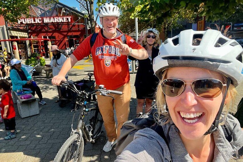 A person on a bicycle with a helmet, another person in a red shirt, and a public market in the background. 1