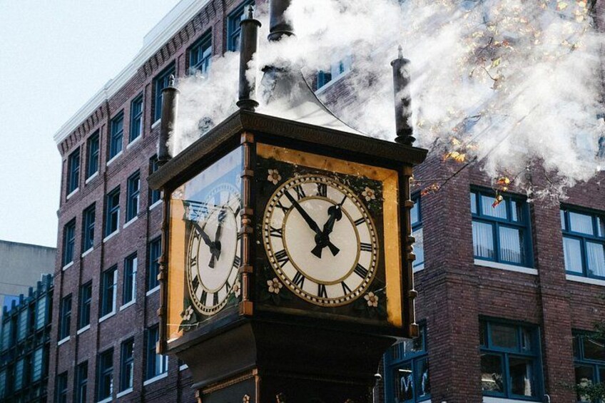 A clock tower with two faces, surrounded by brick buildings and emitting steam. 2