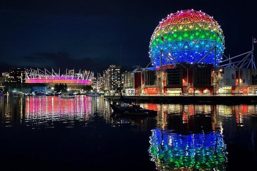 A large, illuminated dome structure reflecting on water, with a brightly lit stadium in the background. 5