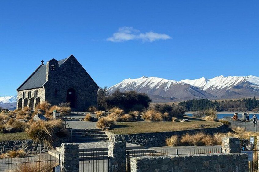 A stone church with a bell tower, surrounded by dry grass and a stone wall, with snow-capped mountains in the background. 2