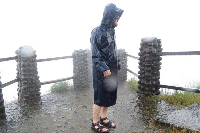 Una persona de pie sobre un puente mojado con barandilla, vestida con un impermeable y sandalias. 1