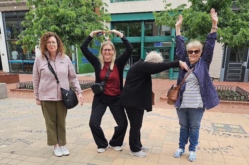 Four people posing with raised arms in an outdoor setting with buildings and trees in the background. 4