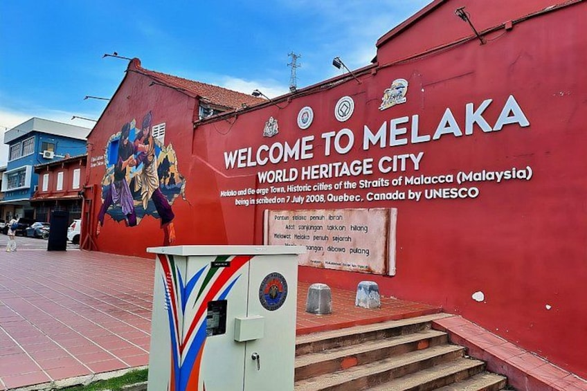 A red wall with a mural and a sign welcoming visitors to Melaka World Heritage City. 1