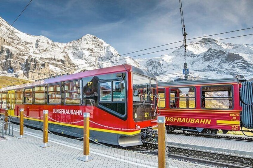 Two red trains labeled "Jungfraubahn" at a station with snow-covered mountains in the background. 1