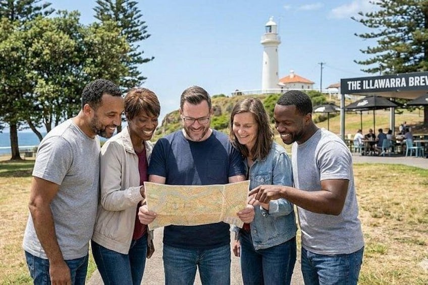 En gruppe mennesker undersøger et kort udendørs, med et fyrtårn og et skilt for 'The Illawarra Brewery' i baggrunden. 1