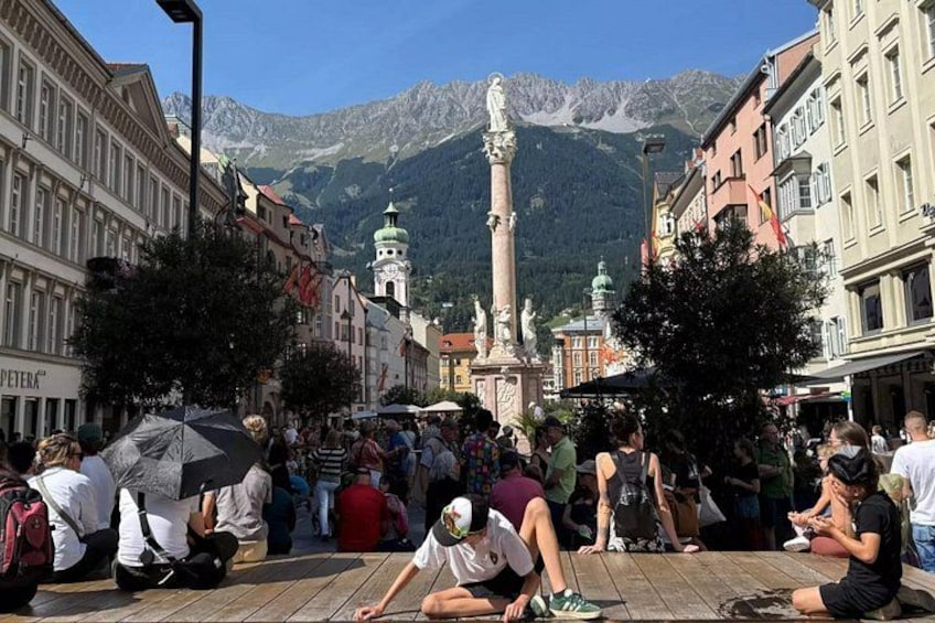A bustling square with people sitting and walking, surrounded by historic buildings and a prominent statue. 5