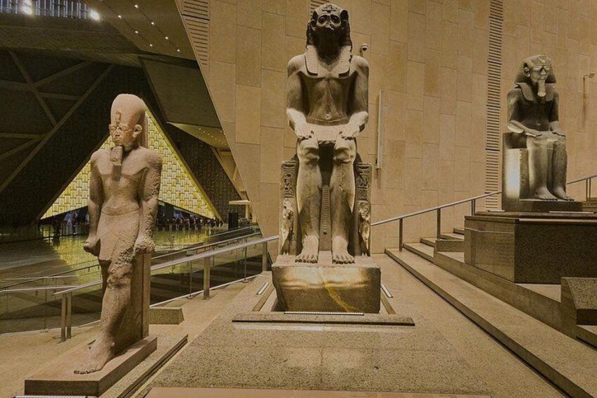 Interior de museo con grandes estatuas de piedra que representan figuras del antiguo Egipto, una estructura en forma de pirámide y una escalera que conduce a una plataforma. 4