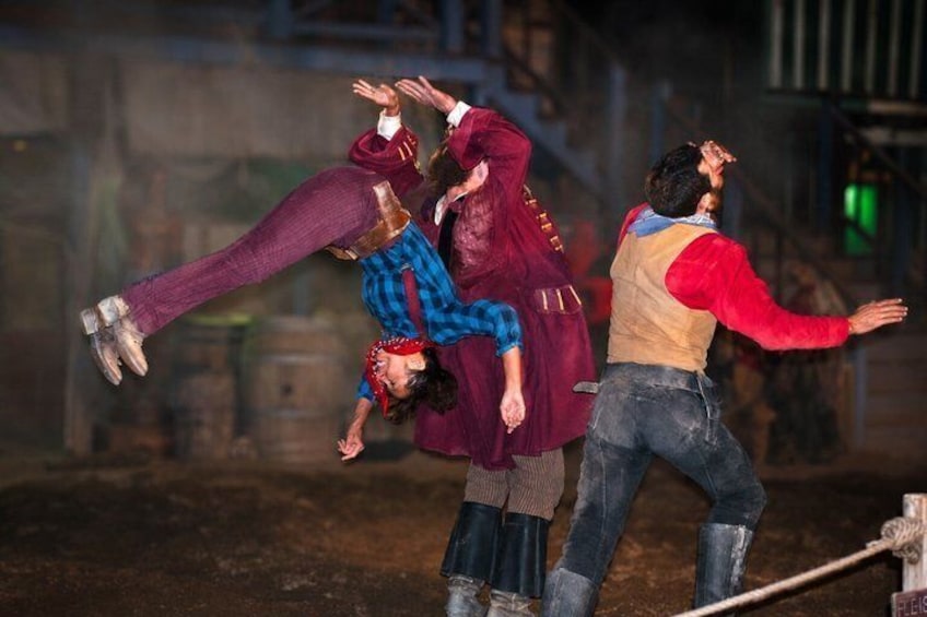 A group of people performing an acrobatic stunt in an industrial setting with metal structures and barrels. 2