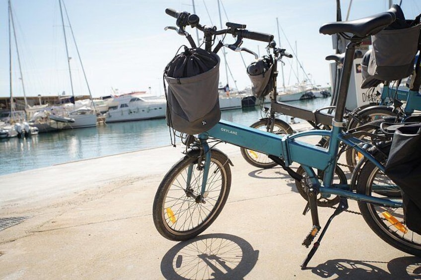 A blue bicycle with a basket and a bag attached, parked near a marina with boats in the background. 3