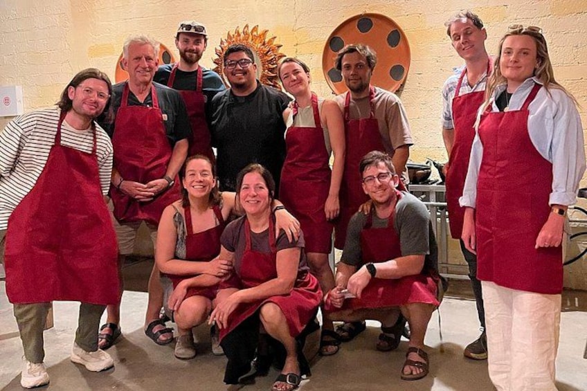 Un groupe de personnes, portant des tabliers et, pour certaines, des chapeaux, se trouve dans une pièce où se dressent une grande table ronde et un mur orné d’un motif circulaire. 2