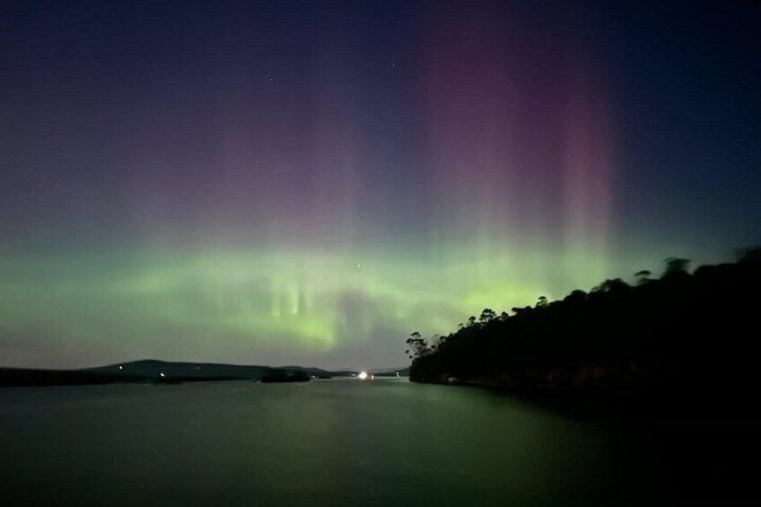 L'aurora boreale riflessa su acque calme, con la silhouette di alberi e luci lontane. 3