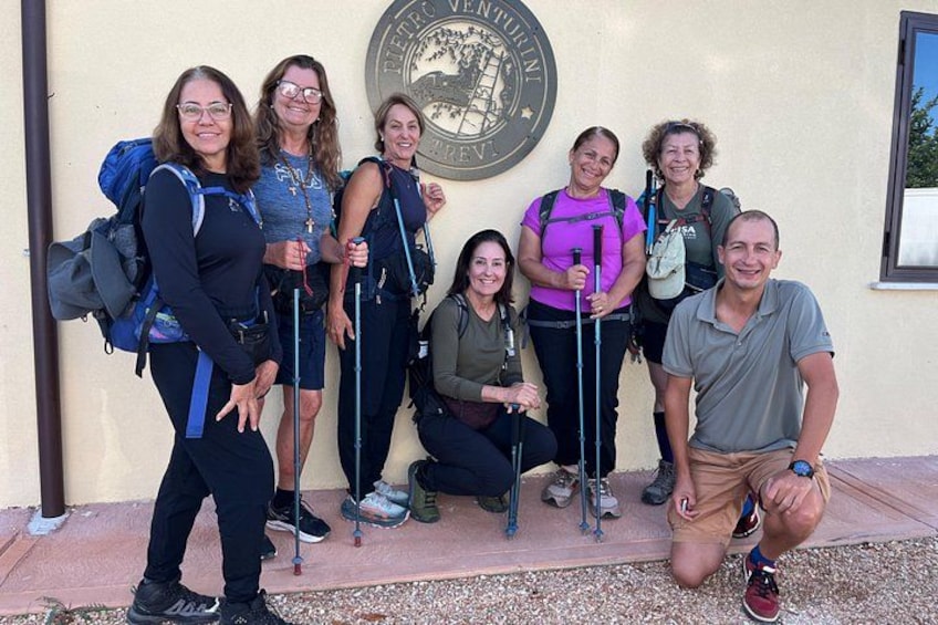 A group of people with backpacks and hiking poles, standing and kneeling in front of a building with a plaque. 2