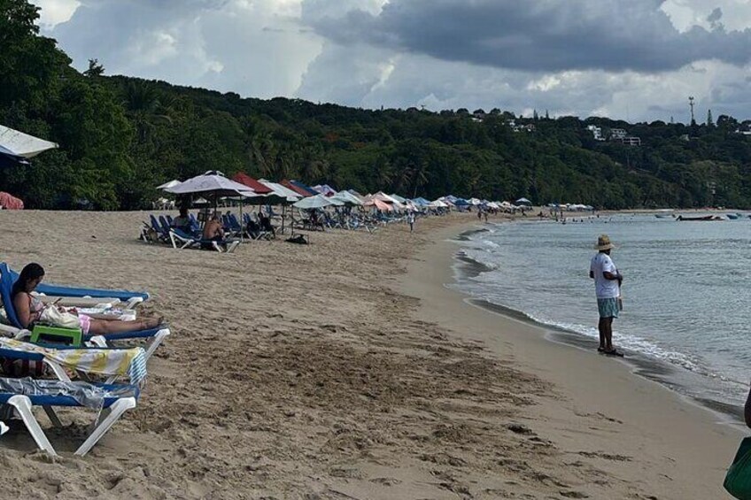 A beach with people relaxing on sun loungers and umbrellas, a person standing by the water, and a forested hill in the background. 3