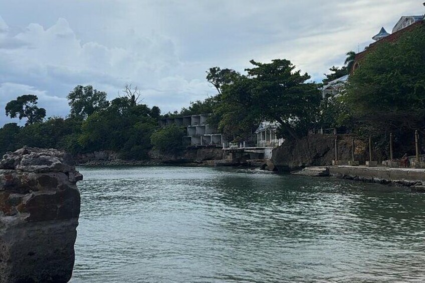 A calm river with a rocky outcrop in the foreground, a bridge in the middle distance, and buildings with trees in the background. 2