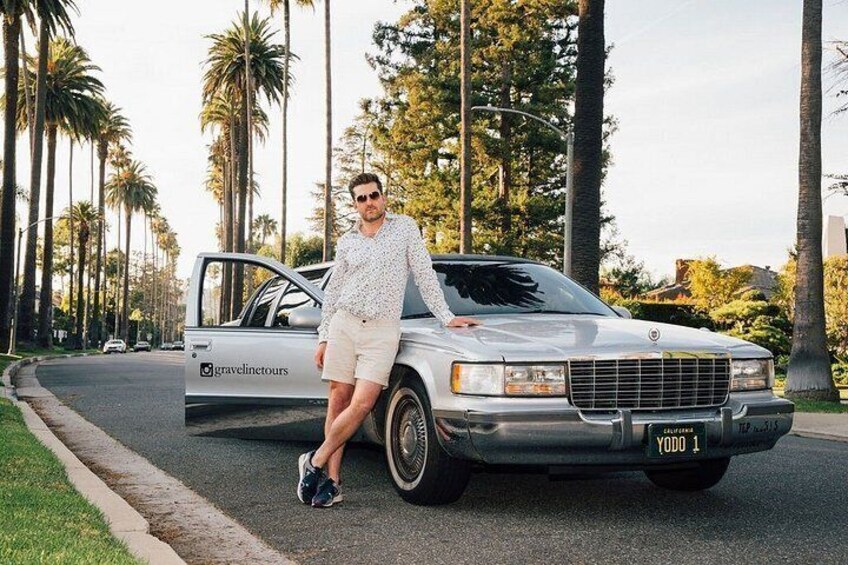 A man standing next to a car with a license plate reading 'YODO 1' on a street lined with palm trees. 2