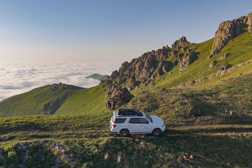 En hvid SUV holder parkeret på en græsbevokset bjergvej med stengrund og en klar himmel. 1