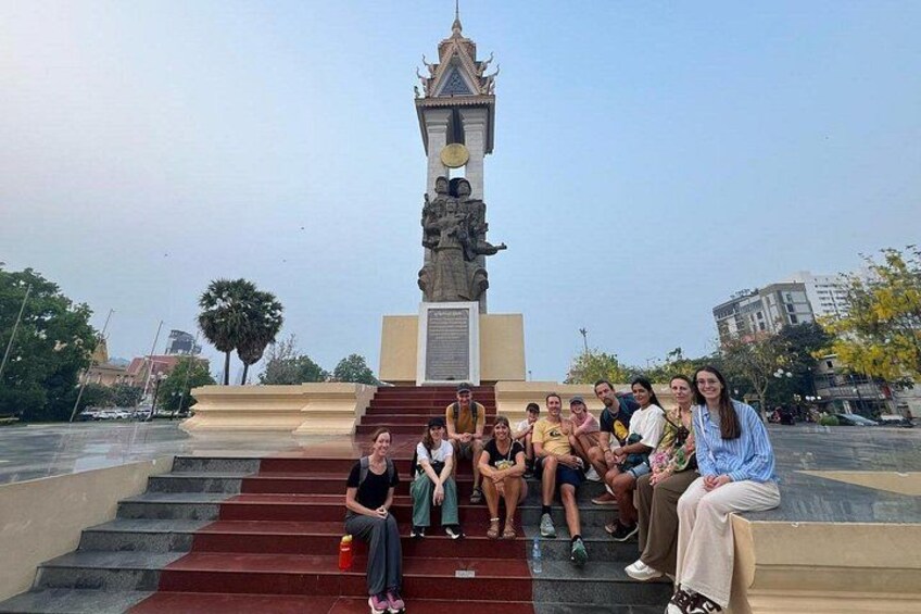 A group of people sitting on steps in front of a monument with a statue on top. 1