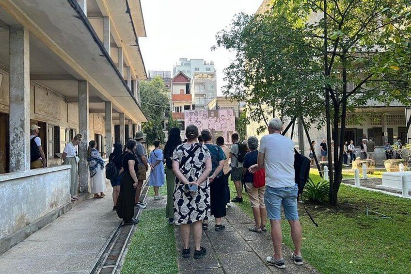 A group of people standing in a courtyard with a building on one side and a tree on the other. 2