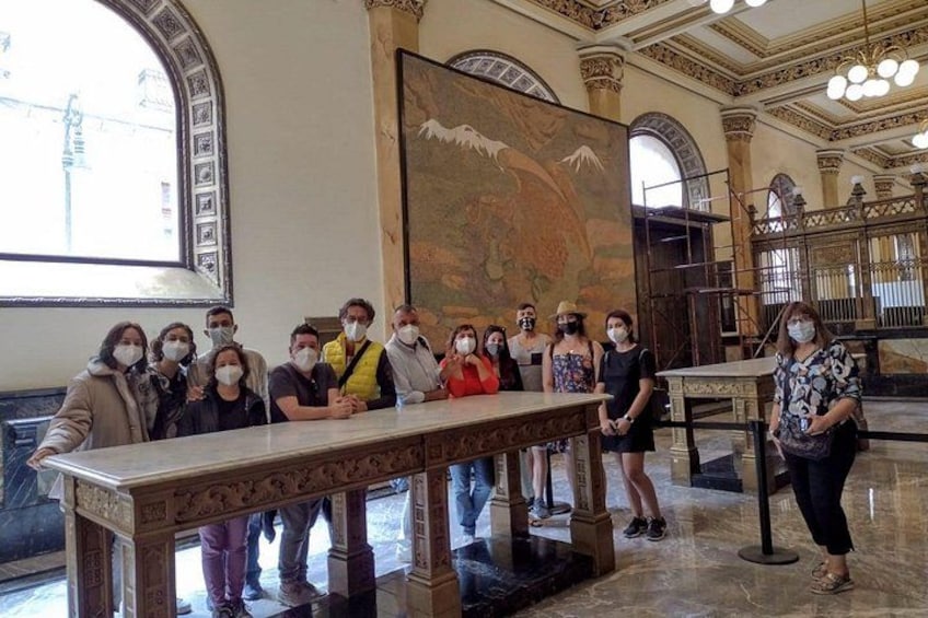 A group of people standing in a grand hall with ornate columns and a large mural on the wall. 2