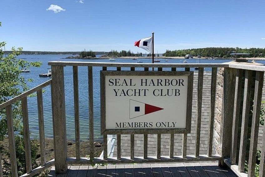 A wooden dock with a sign for Seal Harbor Yacht Club, a flag, and a body of water with boats. 3