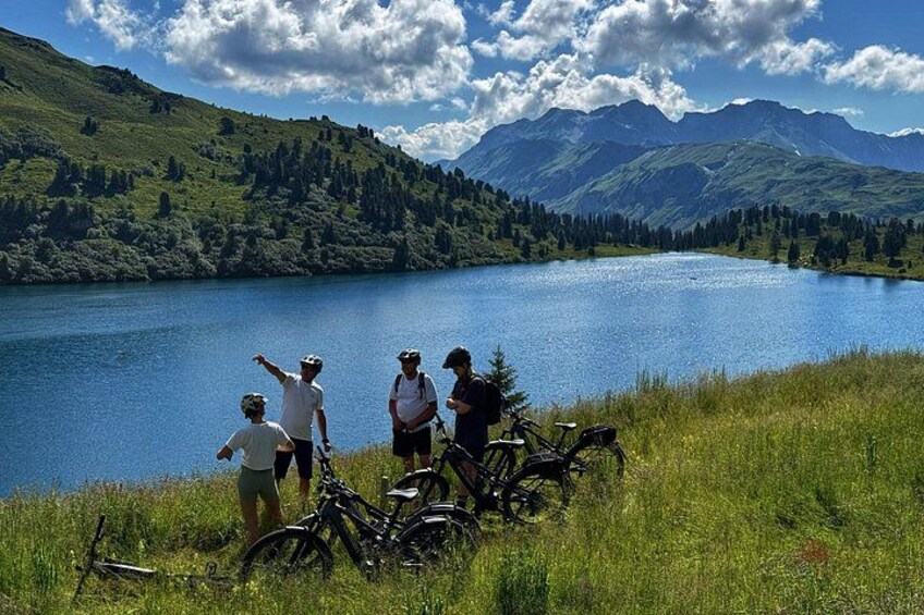 Een groep mensen staat bij een meer, met fietsen, omringd door glooiende heuvels en bergen. 1