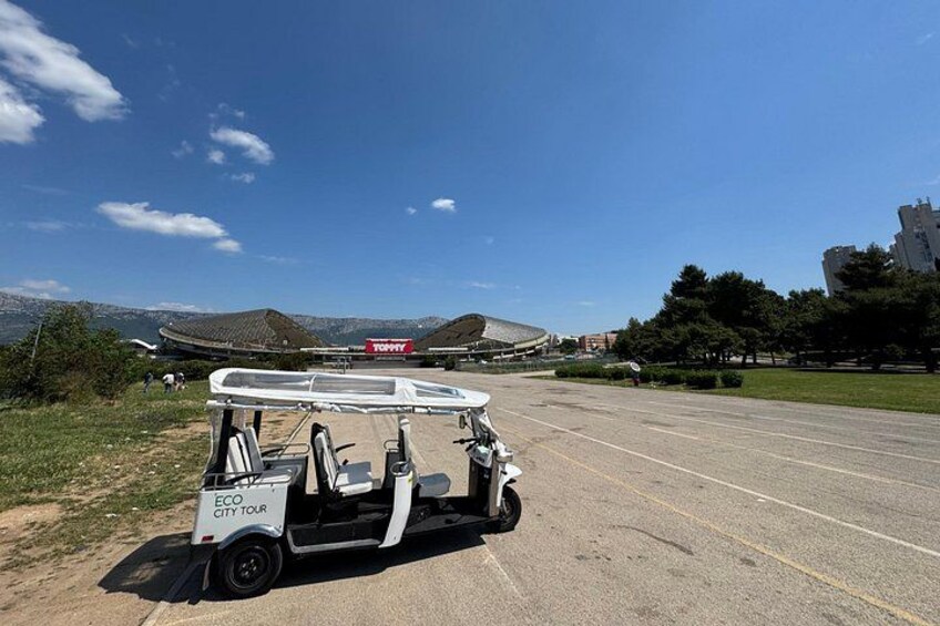 Un golf cart parcheggiato su una strada, con cielo sereno, un edificio in lontananza con la scritta 'TOMMY' e un paesaggio collinare sullo sfondo. 4