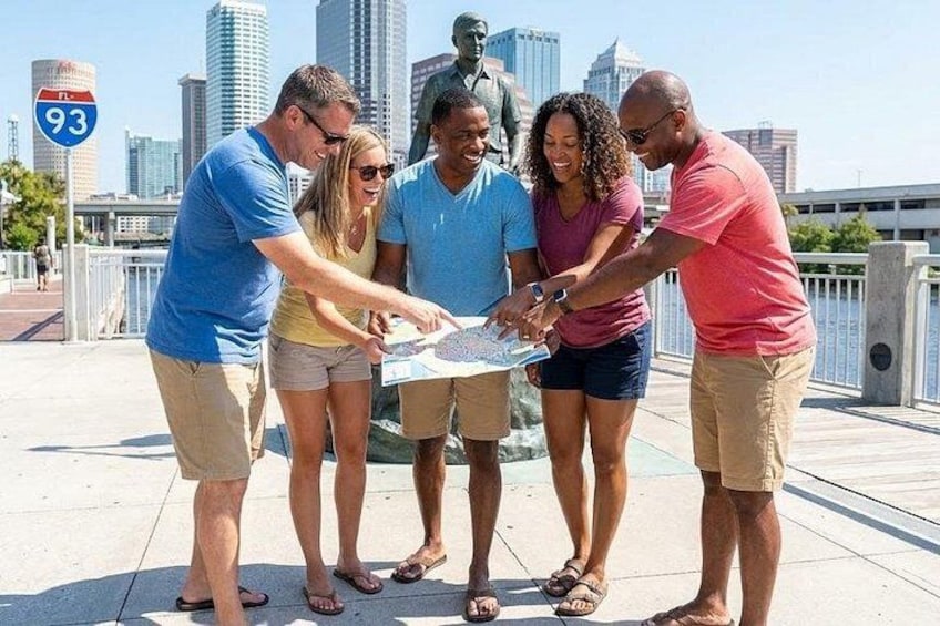 Un grupo de personas revisando un mapa en el paseo marítimo de una ciudad, con una estatua y rascacielos al fondo. 1