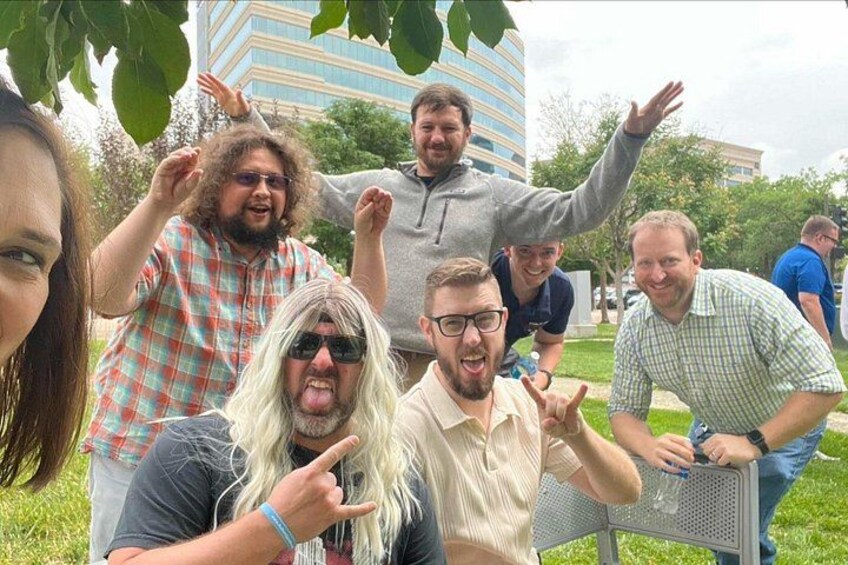 Un grupo de personas al aire libre, algunas sentadas en un banco de metal, otras de pie, con árboles y un edificio al fondo. 3