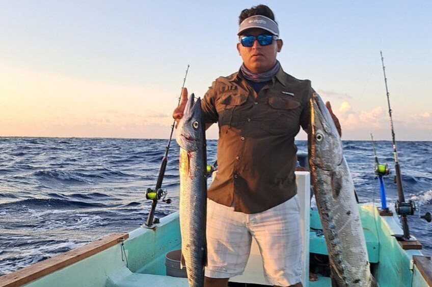 Una persona en un bote sosteniendo dos peces, con cañas de pescar y un mar tranquilo al fondo. 5