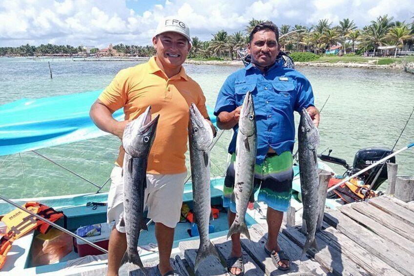Dos hombres sostienen peces en un bote, con un mar azul cristalino y palmeras al fondo. 4