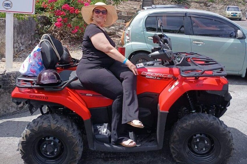 Een persoon zittend op een rode quad met een tas achterop, geparkeerd op een parkeerplaats, met een lichtblauwe auto op de achtergrond. 2