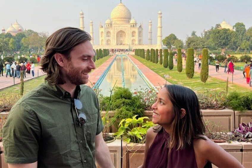 A couple standing in front of the Taj Mahal with a reflection pool and neatly trimmed gardens. 1