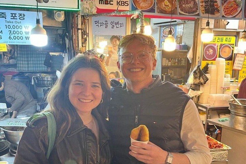 Una persona tiene in mano un cibo in una bancarella, sullo sfondo si vedono diversi cibi e i relativi prezzi. 2