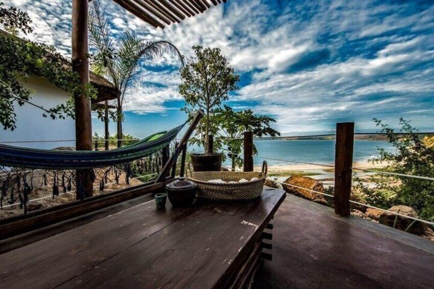 A wooden deck with a hammock, a woven basket, and a potted plant overlooking a beach. 2