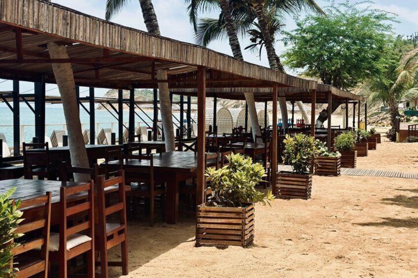 An outdoor restaurant with wooden tables and chairs, surrounded by palm trees and a sandy beach. 3