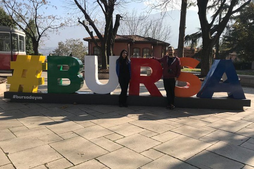 Grandes letras coloridas que deletrean 'BURSA' sobre una superficie pavimentada, con árboles y un edificio al fondo. 3