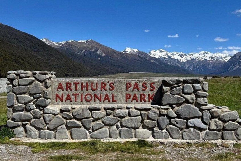 A stone sign with 'ARTHUR'S PASS NATIONAL PARK' written on it, set against a mountainous backdrop. 1