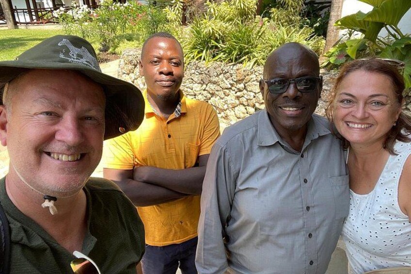 Four people standing outdoors with greenery and a stone wall in the background. 2