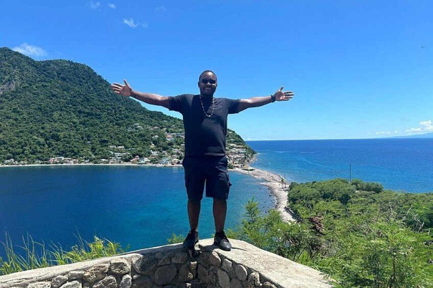 A person standing on a stone platform with arms outstretched, overlooking a coastal landscape with a clear blue sky and a mountainous backdrop. 1