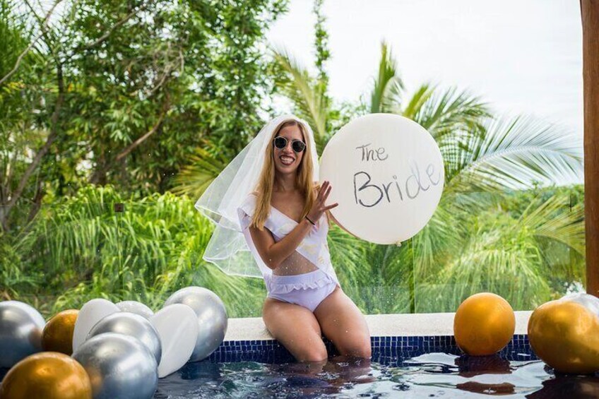 A person in a white dress and veil sitting in a pool surrounded by balloons and greenery. 2