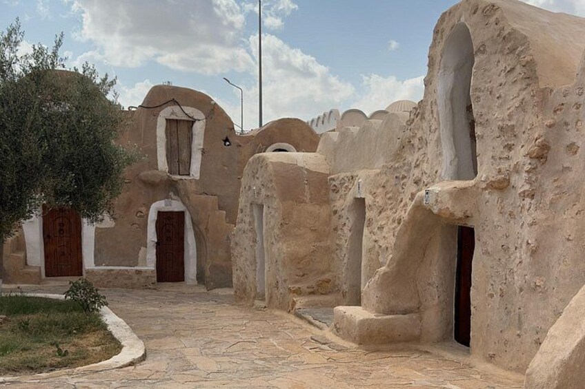 Un pueblo tradicional con construcciones de tierra, un sendero pavimentado y un árbol. 4