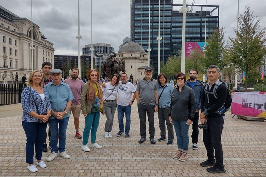 En gruppe mennesker står på et offentligt torv med bygninger og en statue i baggrunden. 5