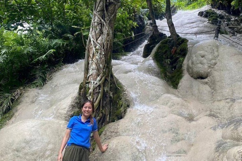 A person standing near a tree by a flowing river with rocks and greenery. 3