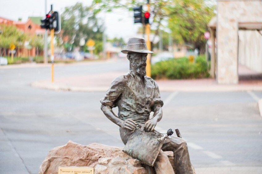 A bronze statue of a man with a hat sitting on a rock at a street corner. 3