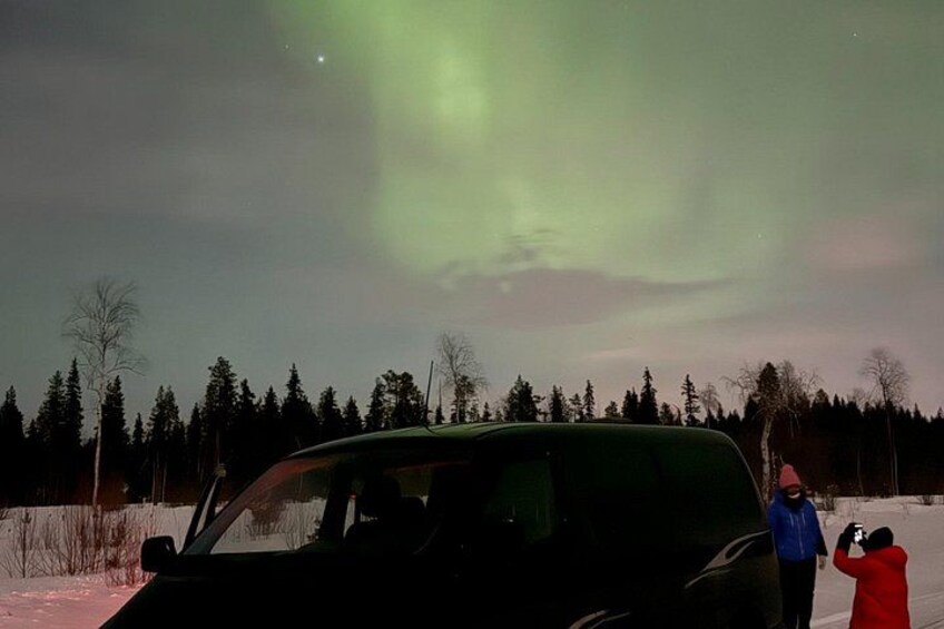 A car, a person in a red coat, and another in a blue jacket observing the aurora borealis. 1