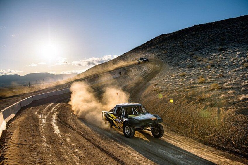 A rally car drifting on a dirt road with dust clouds, surrounded by a hilly landscape and a clear sky. 1