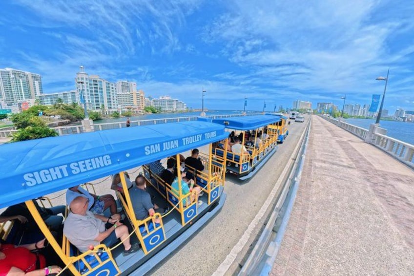 Een trolley in San Juan met passagiers, een brug en stadsgebouwen op de achtergrond. 1